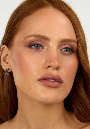 Close-up of a face featuring smooth skin, full matte lips in a nude shade, soft pink eyeshadow, and silver heart-shaped earrings.