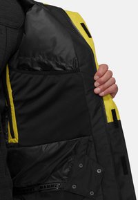 Svart isolerad jacka med insida som har klara gula accenter, flera fickor, mjukt material och dragkedjestängning. MAMMUT-logotyp synlig.