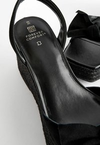 Sorte kilehæl sandaler med en tekstureret vævet platform, blødt ruskindsmateriale, en rund tå og en knyttet sløjfedetalje på stropen.