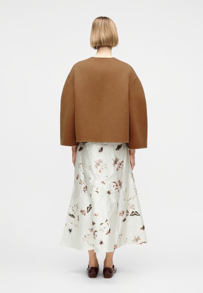 Personne aux cheveux blonds courts portant une grande veste marron et une longue jupe blanche ornée de motifs d'insectes et floraux, vue de dos.