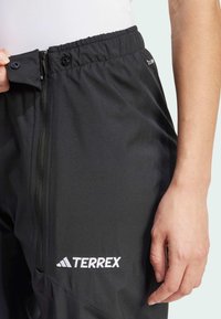 Sorte Terrex bukser med glidelås, elastisk midjebånd og logo-detalj. Materialet ser lett og teksturert ut. Nærbilde av hånd som holder buksene.