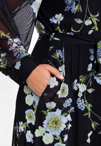 Schwarzes Blumenenkleid mit langen durchsichtigen Ärmeln, das grüne und blaue Blumenmuster aufweist und auf der linken Seite eine Tasche hat. Glatte Stofftextur.