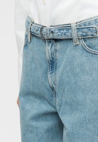 Ανοιχτό μπλε τζιν με μια ταιριαστή ζώνη από τζιν, φορεμένα με ένα λευκό πουκάμισο που έχει μπει μέσα στο παντελόνι, εμφανίζοντας λεπτομέρειες της μέσης μπροστά και της πόρπης της ζώνης.