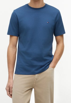 Homme portant un t-shirt bleu uni à manches courtes avec un petit logo sur la poitrine et un pantalon beige, une main dans la poche.