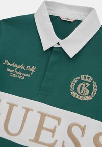 Grøn polo T-shirt lavet af bomuld, med en hvid krave og et stort beige "GUESS" logo. Broderet tekst inkluderer "Los Angeles, Cal." og "1981."