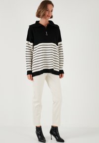 Schwarz-weiß gestreifter oversized Pullover mit Reißverschlusskragen, kombiniert mit cremefarbenen Slim-Fit-Hosen und schwarzen Ankle Boots.
