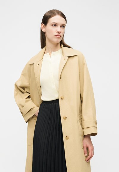 Femme portant un trench-coat beige sur un haut blanc boutonné et une jupe plissée noire, debout devant un fond clair uni.