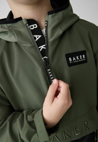 Giacca verde con cappuccio e accenti neri. Presenta una chiusura con zip, un'etichetta con logo e testo in rilievo sul polsino. Tessuto morbido e leggero.