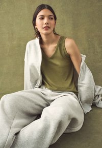 Mladá žena v olivově zeleném tílku a světle šedých teplácích sedí před olivově zbarveným pozadím a dívá se stranou, přičemž má přes ramena přehozenou šedou bundu.