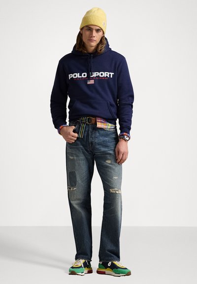 Sudadera azul marino con el texto blanco "POLO SPORT", jeans azules desgastados con parches y zapatillas multicolores; combinada con un gorro amarillo.