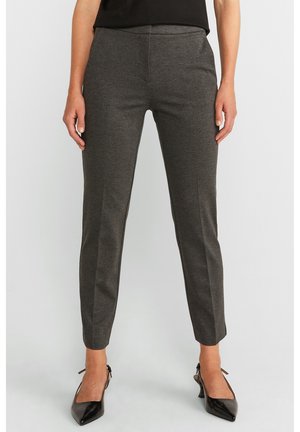 Femme portant un pantalon slim gris foncé longueur cheville et des talons pointus noirs, debout devant un fond blanc uni.