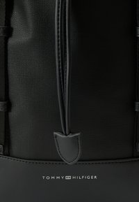 Črn nahrbtnik z zgornjim delom iz strukturirane tkanine, gladkimi usnjenimi detajli in logotipom na dnu. Zapenjanje na zadrgo z izrazitim trakom za vlečenje.