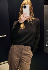 Schwarzer Langarmpullover, kombiniert mit hautfarbenen, strukturierten, hochgerüschten Hosen. Im Hintergrund ist ein Aufzugspaneel mit Tasten zu sehen.
