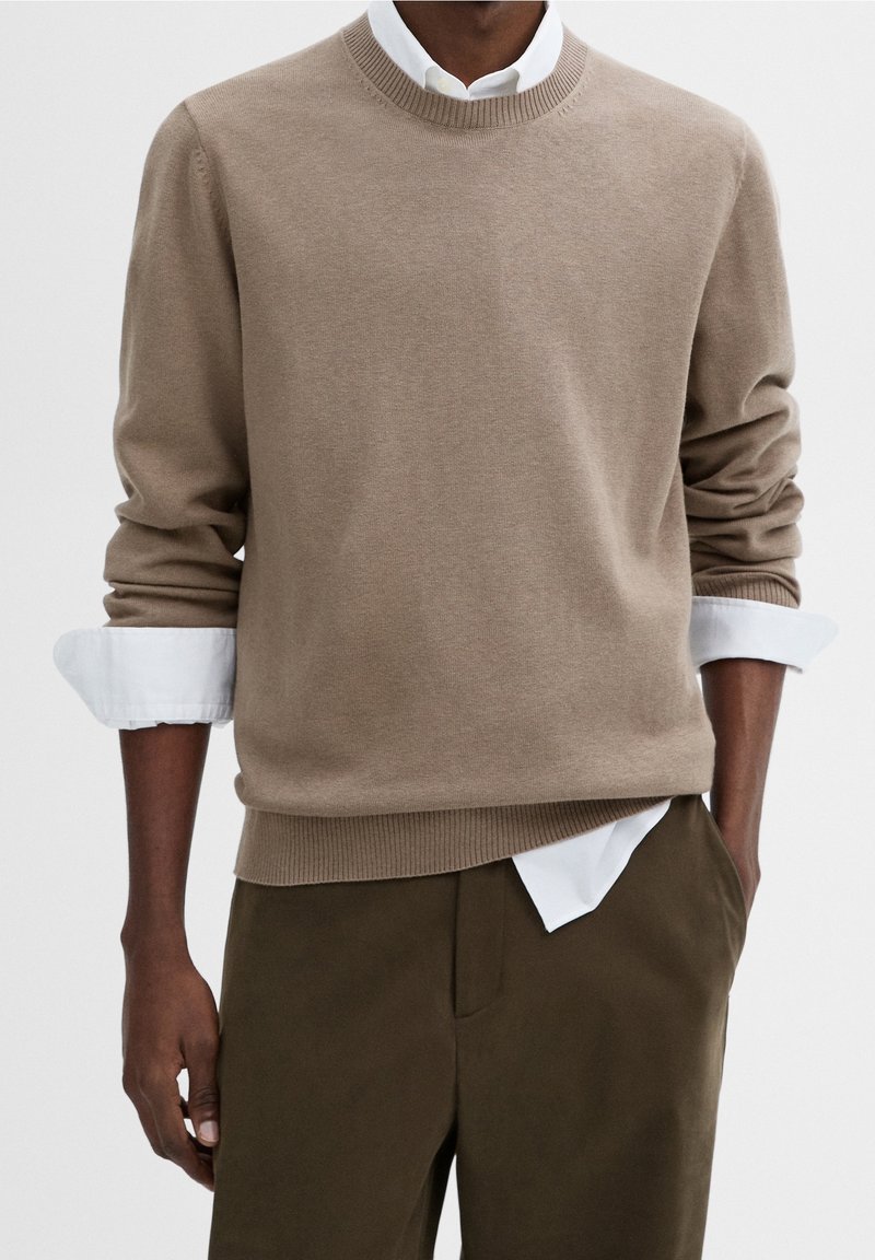 Brauner Strickpullover mit rundem Ausschnitt, langen Ärmeln und gerippten Bündchen. Über einem weißen Hemd mit Kragen getragen und mit olivgrünen Hosen kombiniert.
