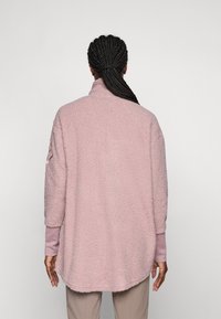 Frau mit geflochtenem Haar, die eine lockere, hellrosa strukturierte Jacke und beige Hosen trägt, von hinten vor einem schlichten Hintergrund gezeigt.