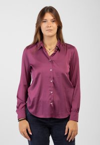 Camicia a maniche lunghe in satin prugna con abbottonatura frontale, orlo arrotondato e texture liscia. Indossata con pantaloni in velluto a coste scuri.