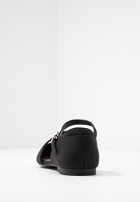 Zwarte platte schoen met een gladde stoffen bovenkant, verstelbare band en een lage, ronde hak. Heeft een minimalistisch ontwerp zonder zichtbare patronen.