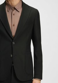 Blazer nero con due bottoni, rever a scollatura, e due tasche frontali, indossato sopra una camicia marrone a trama con colletto a punta.