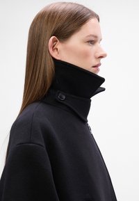 Cappotto di lana nera con colletto alto, dotato di un dettaglio con bottoni al collo e una texture liscia. Vista laterale.