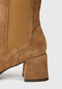 Brown suede ankle boot with a textured fabric side panel, square heel, and subtle stitching details. Smooth and durable finish.