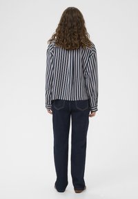 Oseba z kodrastimi rjavimi lasmi, oblečena v srajco z navpičnimi črtami in temnomodre kavbojke, stoji obrnjen z zadnjo stranjo proti beli podlagi.