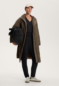 Cappotto lungo oliva trapuntato, top e pantaloni neri, che tiene una borsa a tracolla nera e compatta, con sneakers chiare e un cappellino beige.