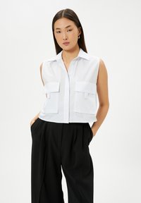 Witte, mouwloze blouse met een knoopsluiting aan de voorkant en twee grote, platte zakken. Gemaakt van gladde stof, gecombineerd met losse zwarte broeken.