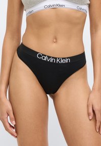 Μαύρο βαμβακερό εσώρουχο σλιπ με φαρδιά ζώνη μέσης που φέρει την επιγραφή «Calvin Klein» με λευκά γράμματα· λεία υφή και σχεδιασμός με ελάχιστη κάλυψη.