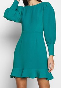 Robe turquoise en tissu texturé, avec un col rond, des manches bouffantes et un ourlet volanté évasé pour plus de détails.