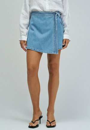 Mujer con falda cruzada de denim azul atada a la cintura, blusa blanca de manga larga y sandalias negras de tacón abiertas, de pie sobre fondo neutro.