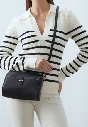 Sort, struktureret håndtaske med guldfarvede metaldele, fastgjort strop og en glat tekstur. Bæres med en hvid og sortstribet sweater.