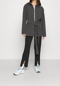 Camisa larga a rayas grises con botones en el frente y cintura ajustable, combinada con pantalones negros con aberturas laterales y zapatillas blancas.