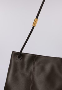 Bolso tote de cuero marrón oscuro con una textura suave, que cuenta con una correa larga con un acento metálico dorado y detalles de costura.