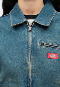 Τζιν μπουφάν με φερμουάρ στο εμπρός μέρος, τσεπάκι στο στήθος με φερμουάρ και κόκκινη ετικέτα της μά ρκας Dickies στο αριστερό στήθος, φορεμένο πάνω από μαύρα και λευκά πουκάμισα.