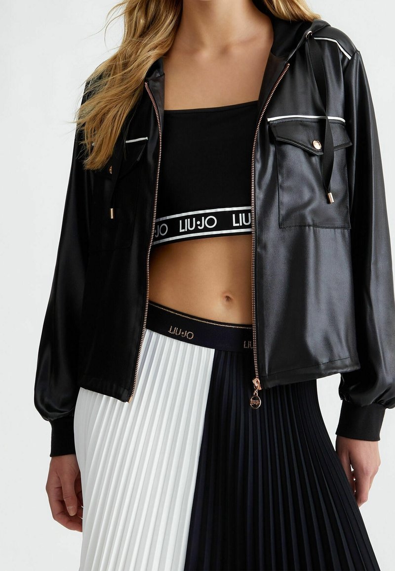 Giacca nera in raso con cerniera, cappuccio, finiture dorate e tasche frontali, indossata sopra un top corto nero aderente e una gonna plissettata bianca e nera.