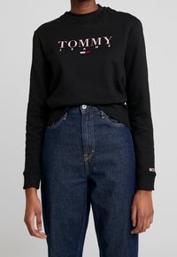 Černá mikina s logem "TOMMY JEANS" v světle růžové. Má žebrovaný límec a manžety, v kombinaci s tmavě modrými džíny.