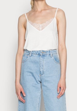 Vrouw draagt een wit cami-topje met kanten randje, ingestopt in hooggehuisde lichtblauwe spijkerbroek, staand met ontspannen armen.