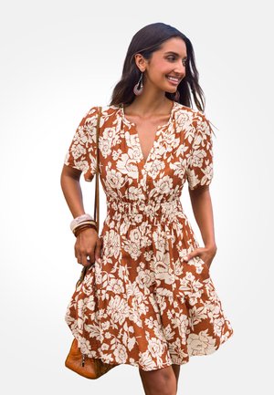 Bruine bloemenjurk met korte pofmouwen, een empire taille en een gelaagde rok. Geaccessoireerd met een kleine bruine handtas en armbanden.