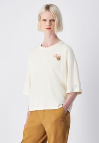 Camiseta de manga corta color crema con cuello redondo, que presenta un estampado de mariposa y logo en el pecho. Tejido de algodón suave, corte holgado.