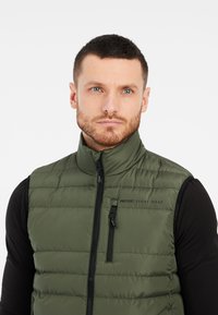 Mann mit kurzen Haaren und Bart, der eine grüne, ärmellose Steppweste mit Reißverschluss über einem schwarzen Langarmshirt trägt und leicht zur Seite schaut.