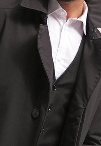 Schwarzer wasserdichter Trenchcoat mit hohem Kragen, Reißverschlussfront und Knopfdetails, über einem weißen Hemd und einer schwarzen Weste getragen.