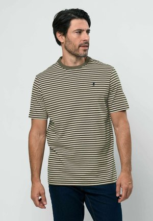 Egy férfi olívazöld és fehér csíkos rövid ujjú pólót és sötétkék farmert visel, és jobbra néz egy egyszerű háttér előtt.