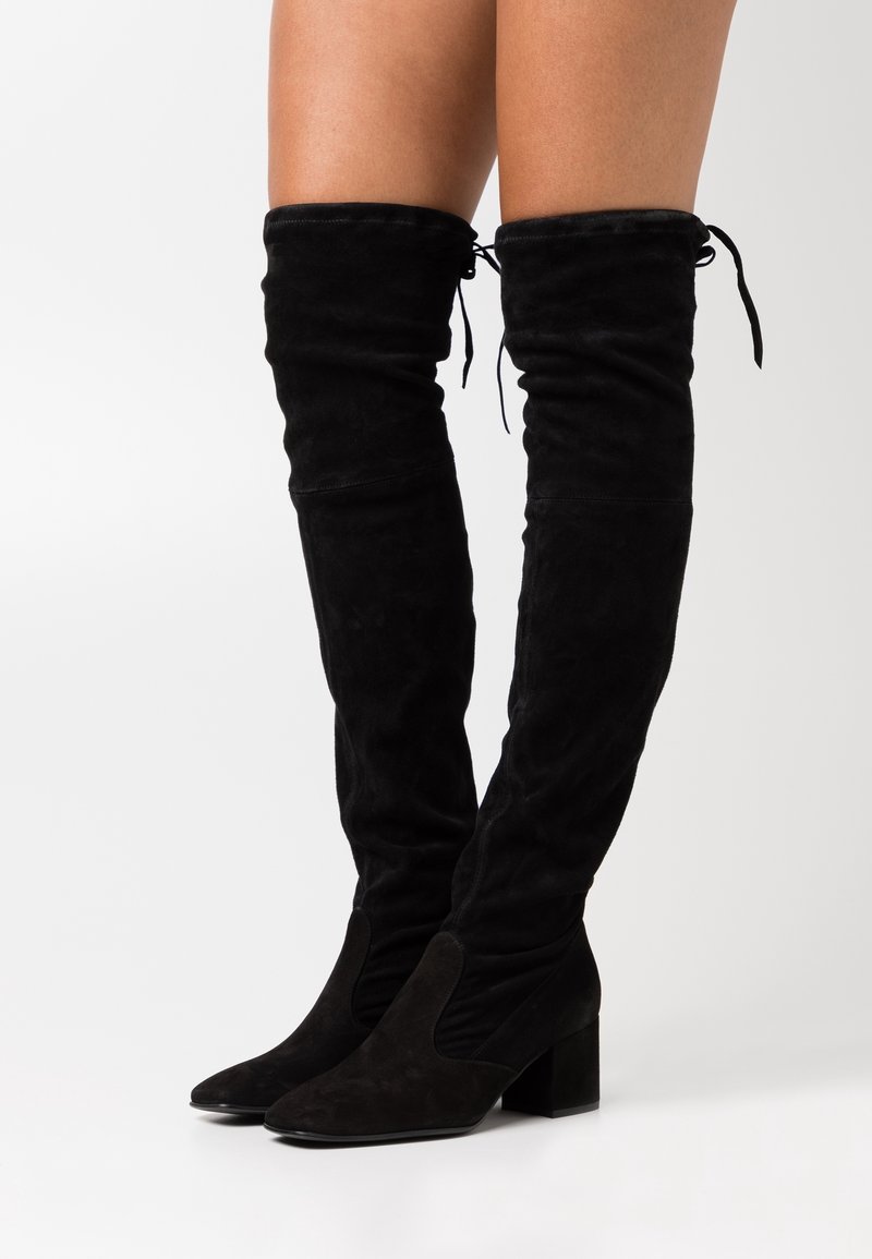 Bottes noires en daim, montantes au-dessus du genou, avec talons carrés, ajustées autour des mollets, et détail de lacets noués en haut.