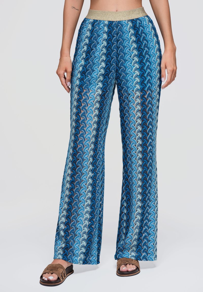 Pantalon large à motifs bleus avec une ceinture élastique dorée, porté avec des sandales plates marron sur une femme debout devant un fond uni.