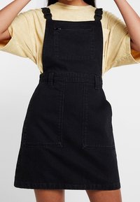 Person som bär en svart denimoverallklänning med luva och ficka fram över en ljusgul kortärmad T-shirt, med armarna uppsträckta.