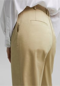 Pantaloni beige con una texture liscia, dotati di tasche laterali, una tasca sul retro e una vestibilità sartoriale. Indossati con una camicia bianca.