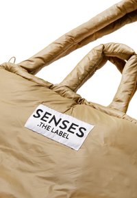 Beige nylon tote bag with padded design, featuring two handles and a rectangular white label with "SENSES .THE LABEL" in bold black text.