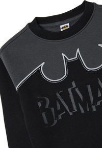 Felpa nera con pannello superiore grigio, caratterizzata da una silhouette di pipistrello e la scritta "BATMAN" in caratteri logori. Collo e polsini a costine.