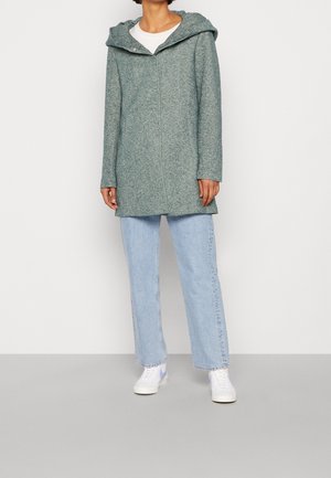 Kobieta ubrana w jasnoniebieskie jeansy o prostych nogawkach, białe trampki, białą koszulę oraz zielony płaszcz z kapturem, stojąca na tle jednolitego jasnego tła.