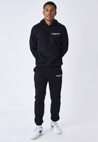 Sudadera negra con el logo "PROJECT X PARIS" y pantalones a juego, hechos de un tejido suave, combinados con zapatillas Nike blancas. Diseño simple y moderno.